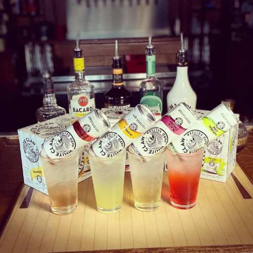A bar scene featuring colorful cocktails in glasses, with various liquor bottles and canned drinks in the background.