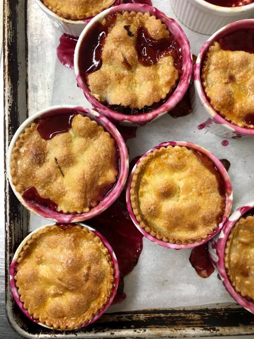 Baked fruit desserts in pink ramekins, with golden crusts and berry filling spilling over, on a baking tray.