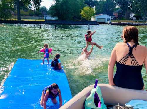 A group of children playing in the water, with one being lifted by an adult while others splash around on a blue mat.