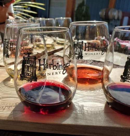 A wooden tray holds several glasses of wine, showcasing different colors, with "Harbinger Winery" logo visible on each glass.