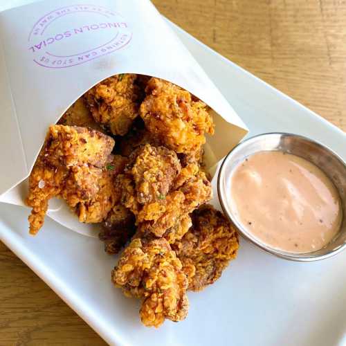 Crispy fried chicken pieces in a paper cone, served with a side of dipping sauce on a white plate.