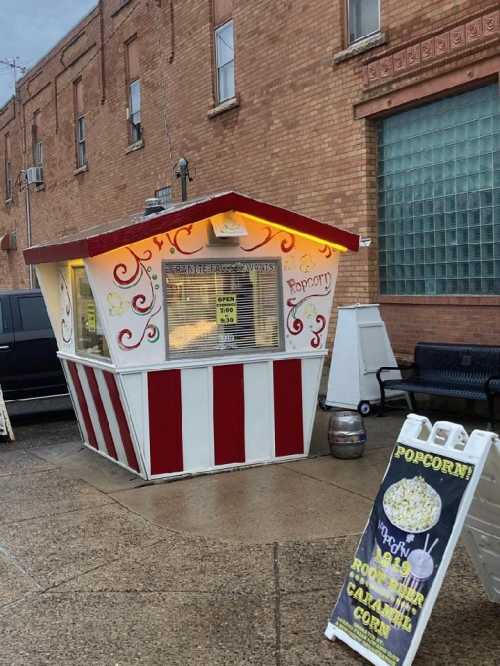 A colorful popcorn stand with a striped exterior, open for business, next to a sign advertising popcorn varieties.