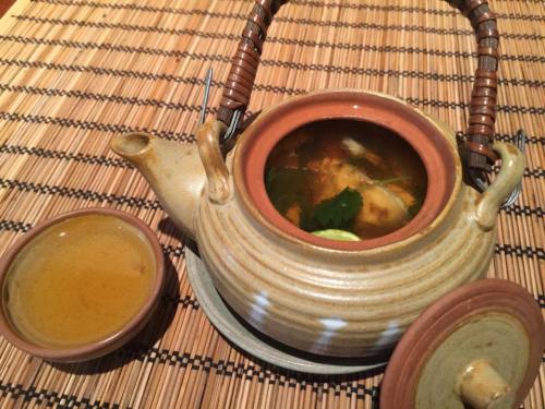A clay teapot with soup and herbs, accompanied by a small bowl of broth, on a woven mat.
