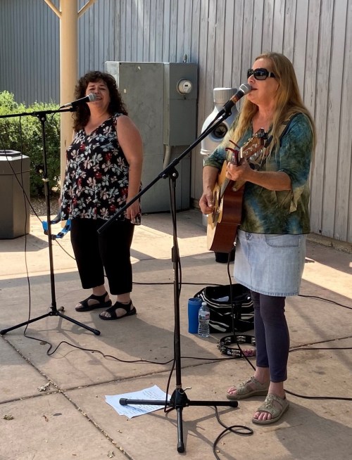Two women perform on stage, one singing into a microphone and the other playing guitar, both enjoying the music.