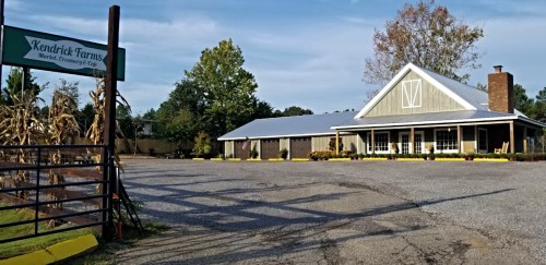 A rustic farm building with a porch, surrounded by trees and a gravel parking area, marked by a sign for Kendrick Farms.