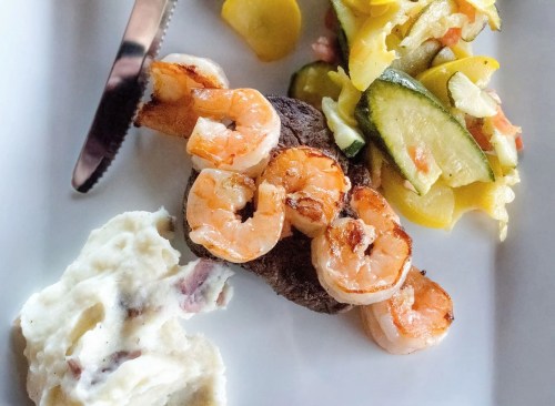 A plate featuring grilled shrimp on a steak, with mashed potatoes and a side of sautéed vegetables.