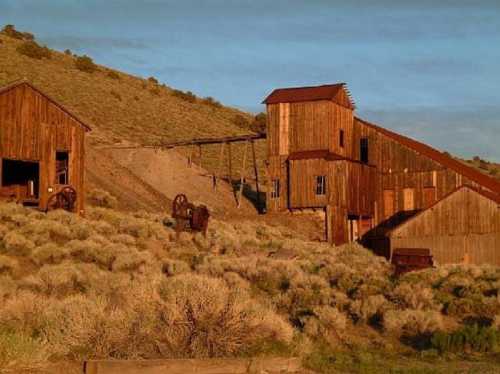 A rustic wooden building complex on a hillside, surrounded by sagebrush and bathed in warm sunlight.