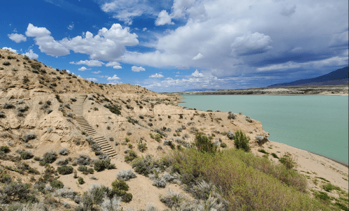 A scenic view of a sandy hillside with stairs leading down to a turquoise lake under a partly cloudy sky.