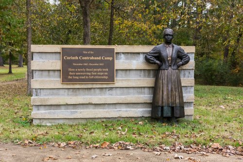 A bronze statue of a woman stands beside a sign marking the site of the Corinth Contraband Camp in a wooded area.