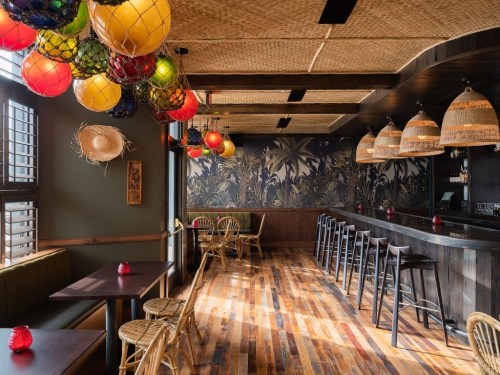 A vibrant bar interior with colorful hanging lights, woven decor, and wooden floors, featuring cozy seating and a tropical mural.