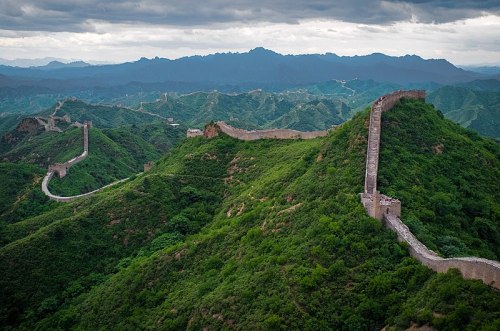 Aerial view of the Great Wall of China winding through lush green mountains under a cloudy sky.