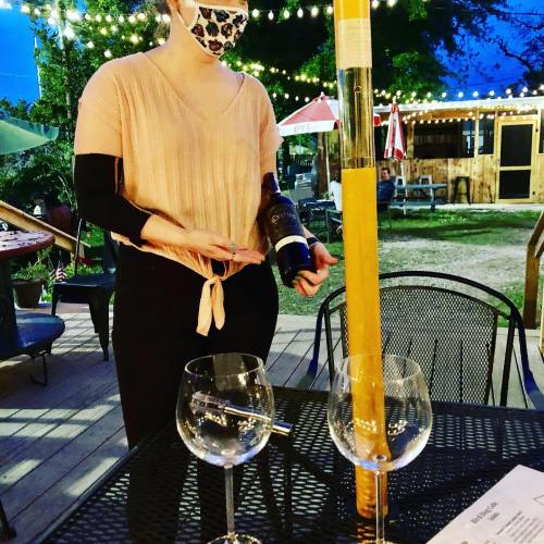 A masked server pours wine into a glass at an outdoor dining area with string lights and tables.