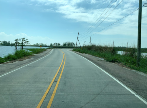 A winding road beside a flooded area, with power lines and greenery under a blue sky.