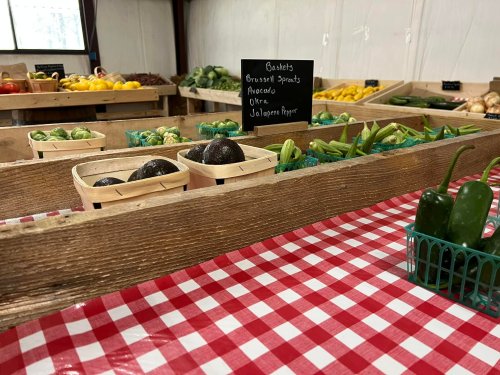 A wooden display of fresh vegetables, including avocados, okra, and jalapeños, on a red and white checkered tablecloth.