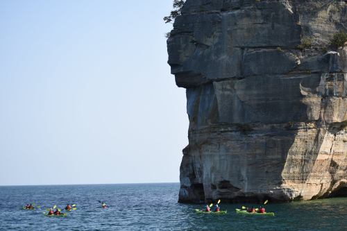 A group of kayakers paddles near a large rock formation by the water on a clear day.