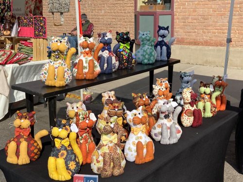 A display of colorful, handmade stuffed cats in various patterns and fabrics, arranged on a table at a market.