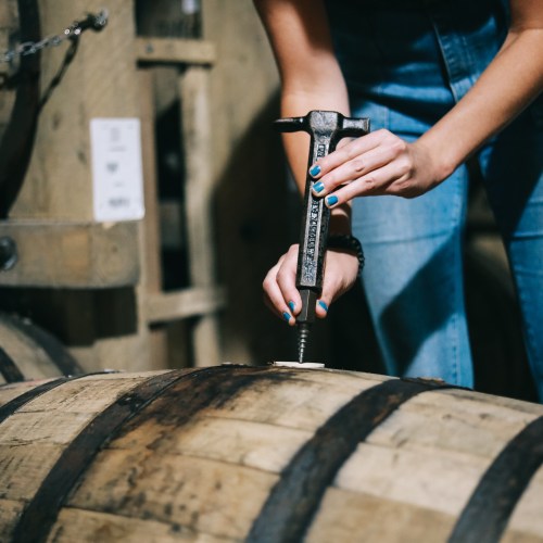 A person in denim overalls uses a tool to inspect a wooden barrel in a rustic setting.