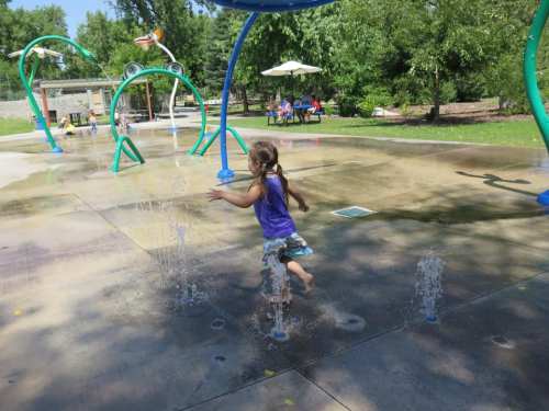 A child in a purple shirt joyfully runs through a splash pad, surrounded by greenery and playful water features.