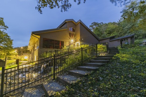 A modern house at dusk, surrounded by trees, with warm lights illuminating the exterior and a staircase leading up.