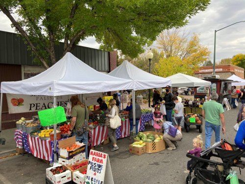 A bustling farmers market with tents selling fruits and vegetables, people shopping, and colorful displays.