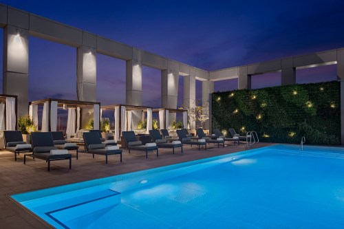 Rooftop pool area at dusk, featuring lounge chairs, cabanas, and a green wall, illuminated by soft lighting.