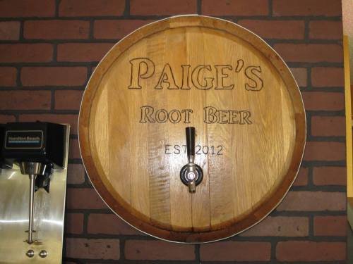 A wooden barrel with "Paige's Root Beer" and "EST 2012" engraved, mounted on a brick wall with a tap.