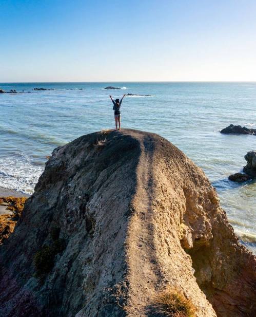 A person stands on a rocky cliff by the ocean, arms raised in celebration, with waves crashing below.