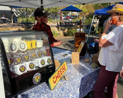 A vendor at a market booth sells cheese, while a customer samples food and enjoys a sunny day outdoors.