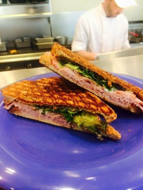 A close-up of a grilled sandwich on a blue plate, filled with ham, lettuce, and pickles, with a chef in the background.