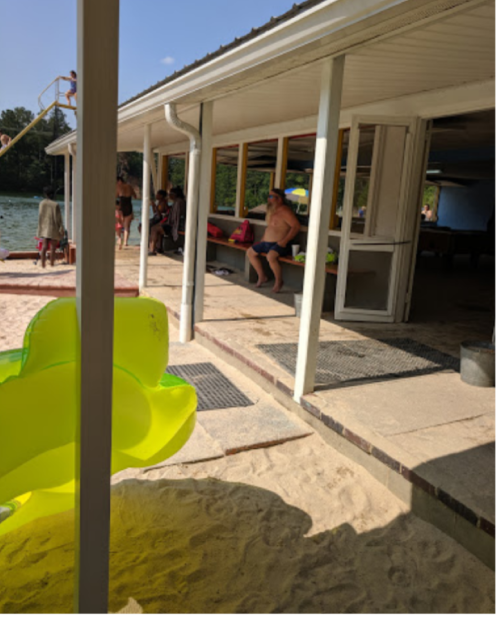 A sunny lakeside scene with a yellow float, sandy area, and people relaxing near a building by the water.