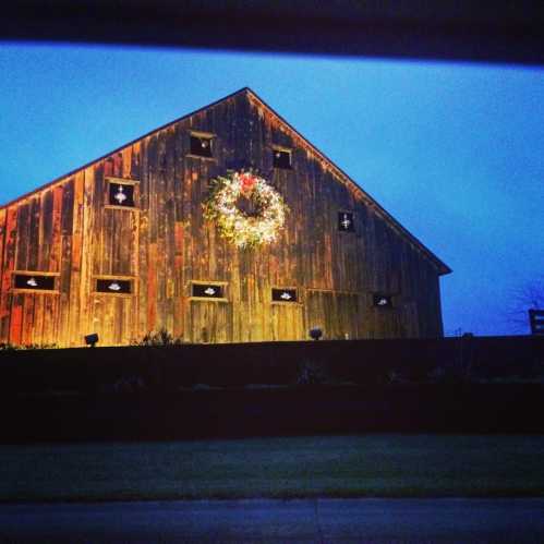 A rustic barn at dusk, adorned with a large wreath on its wooden facade, surrounded by greenery.