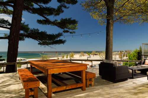 A wooden deck with seating overlooks a beach and lake, framed by trees and string lights against a clear blue sky.