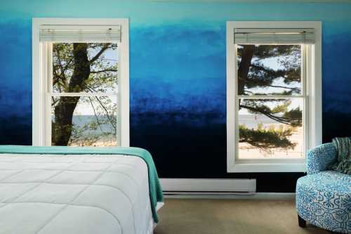 A serene bedroom with a blue ombre wall, two windows showing trees and a beach view, and a cozy chair in the corner.