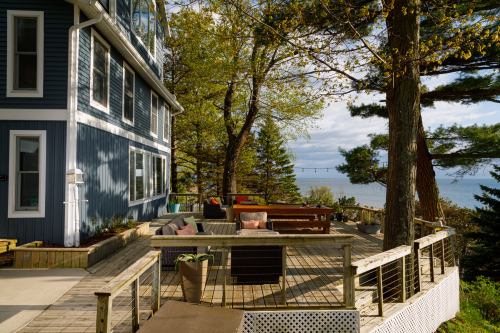 A scenic view of a blue house with a wooden deck overlooking a lake, surrounded by trees and outdoor seating.