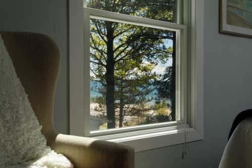 A cozy room with a chair, looking out a window at a tree and a scenic view of water and landscape.