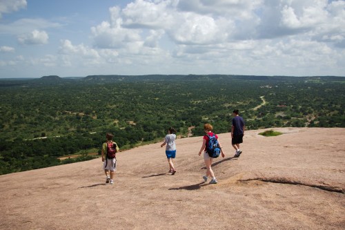 Four hikers walking on a rocky surface, overlooking a lush green landscape under a partly cloudy sky.