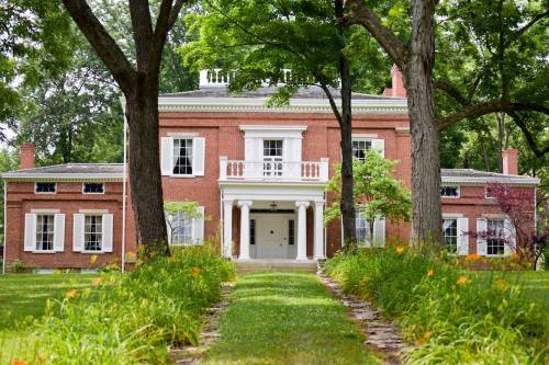 A large brick house with white shutters, surrounded by trees and a garden path lined with flowers.