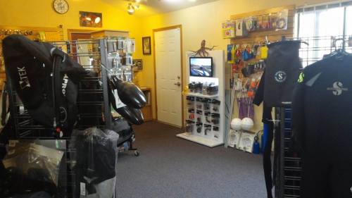 A dive shop interior featuring wetsuits, gear displays, and a monitor, with bright yellow walls and organized equipment.
