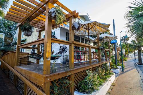 A wooden deck with hanging plants, hammocks, and umbrellas, surrounded by greenery and a pathway.