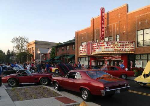 A classic car show outside a vintage theater, with colorful cars parked and a crowd enjoying the event.