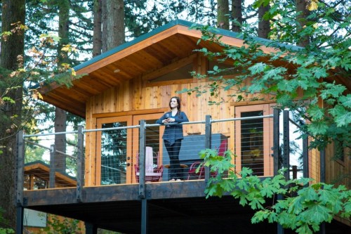 A woman stands on the deck of a wooden cabin surrounded by tall trees, enjoying the serene forest view.