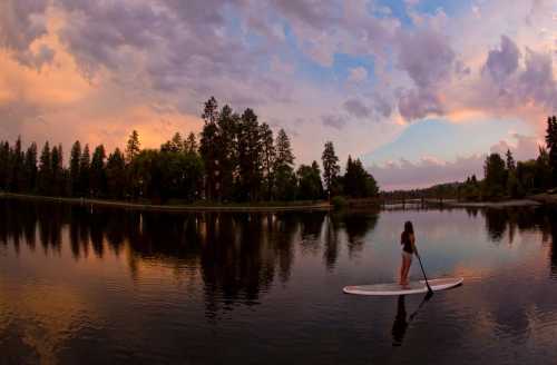 A person stands on a paddleboard in a calm lake at sunset, surrounded by trees and colorful clouds.