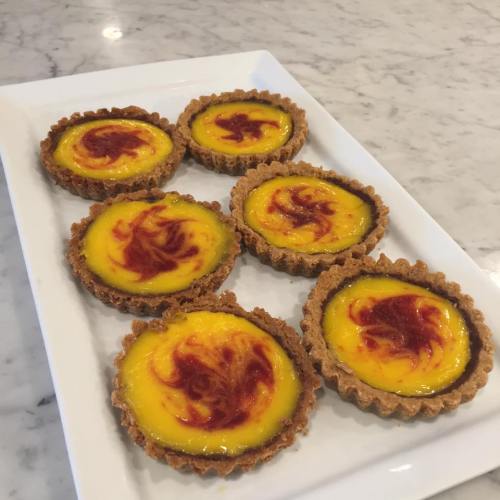 A plate of six golden egg tarts with a swirl of red topping on a marble surface.