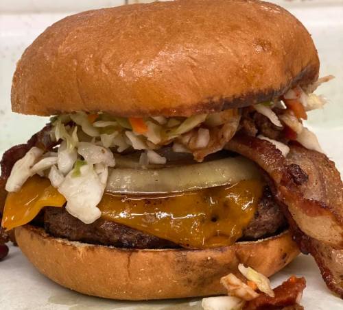 A close-up of a burger with a beef patty, cheddar cheese, bacon, pickles, and coleslaw on a toasted bun.