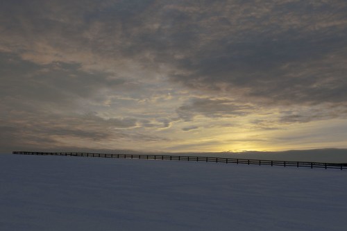 A snowy landscape under a cloudy sky, with a wooden fence stretching across the horizon as the sun sets.