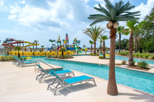 A sunny pool area with lounge chairs, palm trees, and a colorful water park in the background.