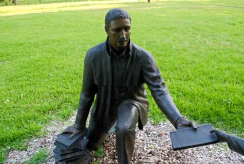 A bronze statue of a man kneeling, holding a tablet, surrounded by green grass and trees.