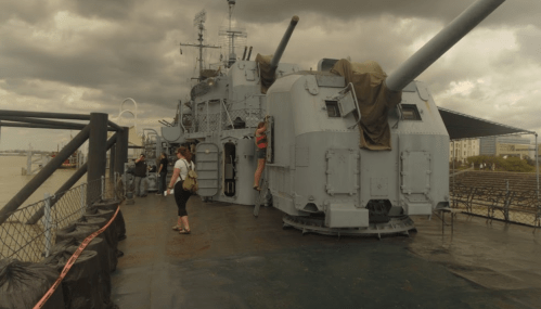 A large naval gun on display at a waterfront, with visitors exploring the area under a cloudy sky.