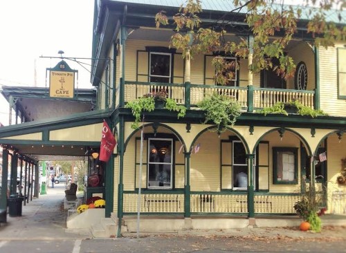 A charming yellow café with green trim, featuring a porch and decorative pumpkins, set in a quaint street.