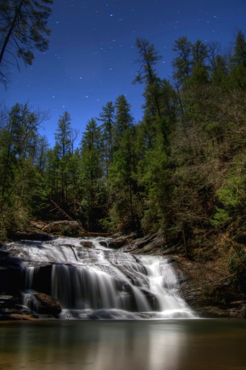 A serene waterfall cascades over rocks, surrounded by lush trees under a starry night sky.